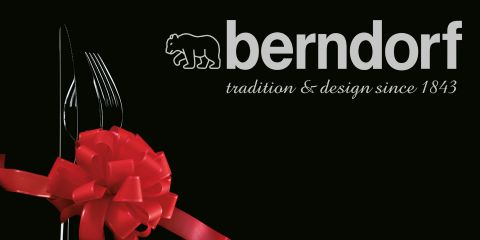 Un juego de cubiertos, que consta de tenedor, cuchillo y cuchara, está atado con un lazo rojo. El texto dice "berndorf tradition & design since 1843", con un logotipo de oso estilizado sobre "berndorf". El fondo es negro.