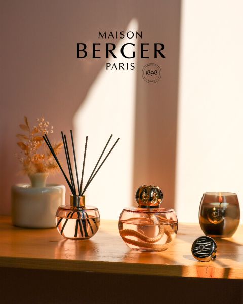 En una estantería soleada se encuentran dos difusores de Maison Berger Paris con un elegante diseño de vidrio. Un difusor está equipado con varillas de ratán, el otro con una tapa decorativa. Junto a ellos hay un pequeño jarrón con flores secas y una vela, creando un ambiente cálido y acogedor.