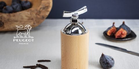 Un molinillo de pimienta de madera y acero inoxidable está al lado de higos enteros y cortados sobre un mantel. Al lado hay un cuchillo. En la esquina superior izquierda se ve el logotipo de Peugeot con un león. En el fondo se ve un cuenco de madera con cápsulas de especias.