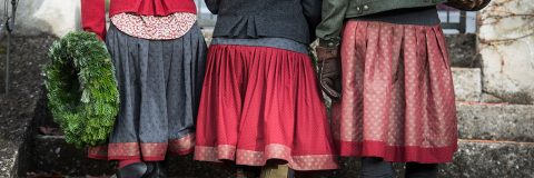Tres personas vestidas festivamente están de pie una al lado de la otra sosteniendo una corona verde en sus manos. Llevan faldas estampadas en tonos rojos y gris oscuro, chaquetas abrigadas y guantes. La escena parece tener lugar al aire libre sobre escalones de piedra.