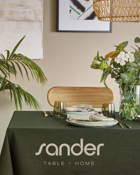 Una mesa de comedor moderna con un mantel verde, platos, vasos, servilletas, un arreglo floral y la inscripción «sander TABLE + HOME».
