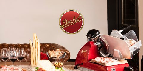 Sobre una mesa con varias carnes, quesos, grissini y copas de vino, se encuentra una antigua cortadora de fiambres para carne roja. Al fondo se puede ver un sofá de cuero y un logo de "Berkel". El ambiente es acogedor y rústico.