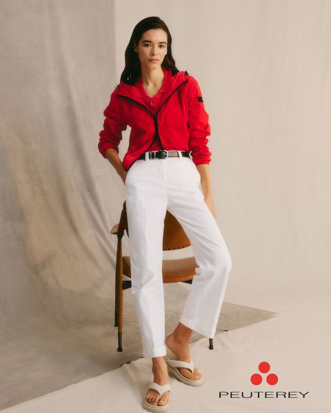 Una mujer con una chaqueta roja y pantalones blancos está apoyada en una silla. Tiene el pelo largo y oscuro y lleva sandalias blancas. El telón de fondo es minimalista con un fondo neutro. El logo de "Peuterey" está en la esquina inferior derecha.