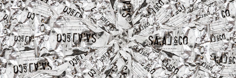 Un montón de envoltorios arrugados de papel de aluminio. Cada pieza tiene un texto impreso que contiene partes de palabras y números como "SALISCO" y "031550". La superficie brillante crea un patrón texturizado y reflectante.