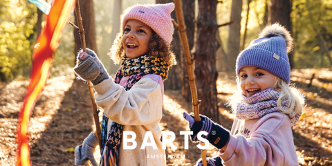Dos niños corren por un bosque iluminado por el sol. Llevan gorros de invierno, bufandas y guantes. Sonríen y sostienen palos con cintas de colores. En la parte inferior de la imagen se ve el texto "BARTS AMSTERDAM".