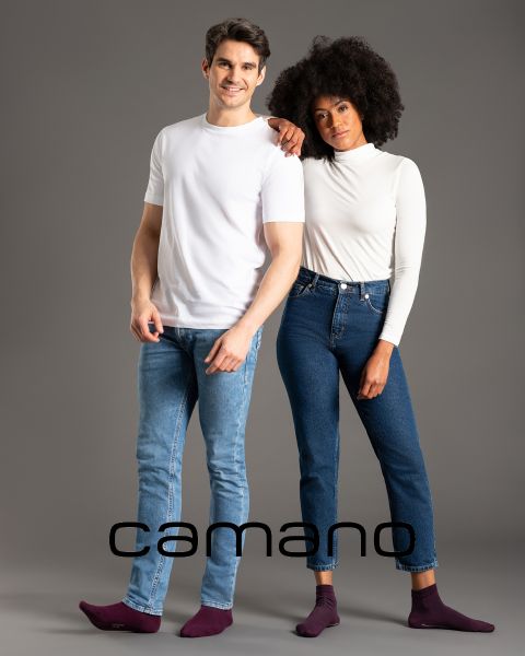 Un hombre con camiseta blanca y vaqueros y una mujer con una blusa blanca de manga larga y vaqueros posan con los brazos entrelazados. Ambos están descalzos. En la parte inferior de la imagen está la palabra "camano".
