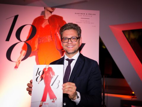 Ein Mann in Anzug und Krawatte lächelt und hält eine Zeitschrift in der Hand, auf der eine Frau in einem roten Outfit abgebildet ist. Die Zeitschrift trägt den Titel „10/13“. Im Hintergrund ist ein großes Poster mit demselben Zeitschriftencover zu sehen. Die Umgebung ist warm beleuchtet.