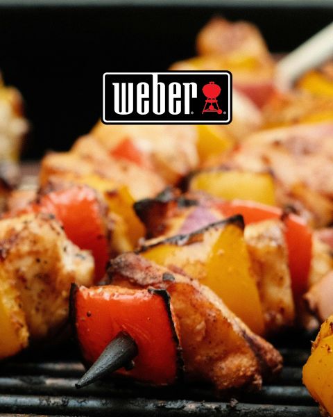 Primer plano de brochetas de pollo y verduras a la parrilla en una barbacoa. Las brochetas están hechas con trozos de pollo con pimientos rojos y amarillos, así como cebollas rojas. El logotipo de Weber se ve prominentemente en la parte superior central de la imagen.