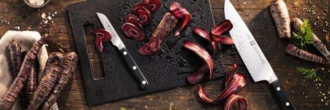 Una mesa de madera oscura con una tabla de cortar negra sobre la que descansan dos cuchillos y zanahorias moradas cortadas. Zanahorias enteras y hojas de zanahoria esparcidas alrededor contribuyen a una escena de cocina rústica.