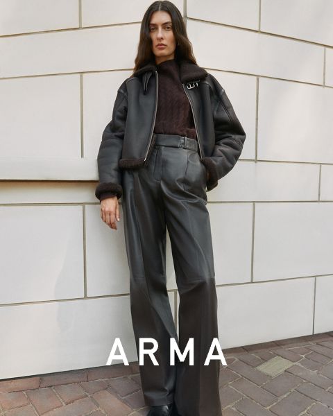 Una persona con pelo largo está de pie frente a una pared alicatada. Lleva una chaqueta negra y pantalones oscuros de talle alto. Tiene la mano izquierda en el bolsillo y en la parte inferior de la imagen aparece la palabra "ARMA".