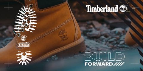 Primer plano de una bota Timberland sobre suelo rocoso, con el logotipo de Timberland y la frase "BUILD FORWARD" en el lado derecho. La bota es de color marrón claro y tiene una suela robusta, que irradia un aspecto de aventura y aire libre.