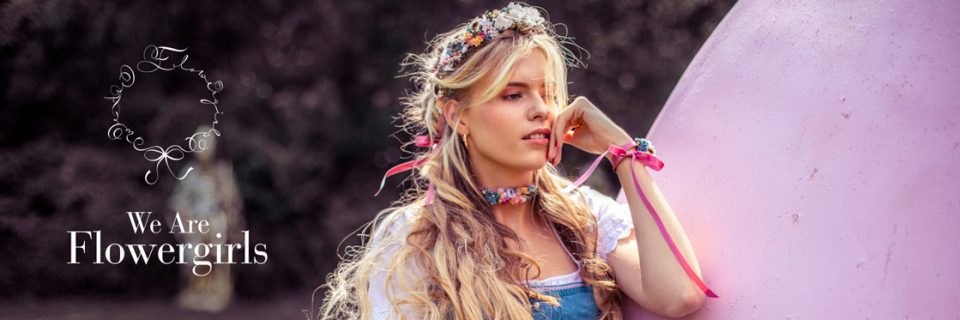 Una mujer con cabello largo y rubio y una corona de flores se apoya en un gran objeto rosa. Lleva un vestido dirndl con un collar de flores y pulseras de cintas. El texto dice "We Are Flowergirls" con un logo floral circular adjunto.