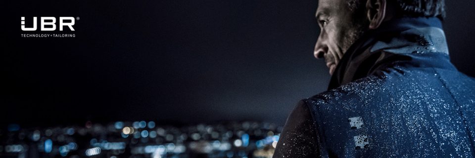 Una persona con una chaqueta mira por encima del hombro un paisaje urbano nocturno. Hay gotas de agua en la chaqueta que reflejan las luces de la ciudad. En la esquina superior izquierda se ven el logotipo de UBR y las palabras "Technology Tailoring".