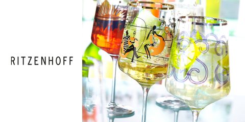 Un conjunto de coloridas y artísticas copas de Ritzenhoff, llenas de bebidas. Cada copa tiene un diseño único con patrones y figuras vibrantes. El fondo muestra reflejos de luz y más copas, creando un ambiente festivo.