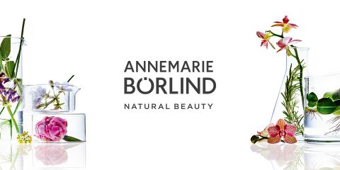 Recipiente de vidrio con flores y hierbas alrededor, que resaltan elementos naturales. En el centro se lee "ANNEMARIE BÖRLIND BELLEZA NATURAL". El fondo es blanco.