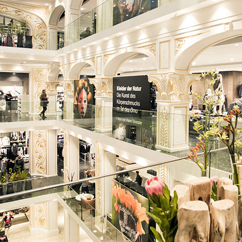 Un centro comercial de varios pisos con interiores intrincadamente decorados en blanco y dorado. Compradores exploran tiendas en diferentes pisos. Se ven grandes carteles y arreglos florales. Barandillas de vidrio y arcos decorativos añaden elegancia a la escena.