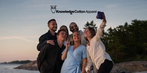 Un grupo de cinco personas se toma un selfie al aire libre cerca del agua, con árboles y una puesta de sol de fondo. Sonríen y posan juguetonamente, con ropa informal. Encima de ellos se ve el texto "KnowledgeCotton Apparel" y un logo de búho.