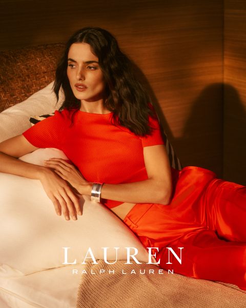 Una mujer con cabello largo y ondulado se reclina en un sofá blanco, vistiendo una blusa roja brillante y pantalones a juego. La cálida luz del sol proyecta sombras sobre la escena. En la parte inferior se lee "LAUREN RALPH LAUREN".