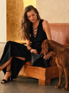 Una mujer con un vestido negro está sentada con un perro marrón en un sofá marrón y sostiene un bolso negro en su regazo.