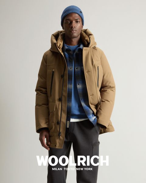 Una persona con un abrigo de invierno marrón claro, un gorro de punto azul y una camisa de cuadros azules sobre una sudadera con capucha está de pie frente a un fondo liso. Debajo aparece la palabra "Woolrich" junto con las ciudades de Milán, Tokio y Nueva York.