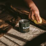 Una persona aplica cera a una prenda marrón con una esponja amarilla. Una lata que dice "Barbour Wax Thornproof Dressing" está cerca sobre una superficie cubierta de tela. La escena está iluminada cálidamente y las sombras sugieren una fuente de luz natural.