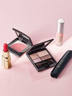 Una serie de productos de maquillaje, que incluyen lápiz labial, rubor, paleta de sombras de ojos, rímel y base, dispuestos sobre una superficie rosa.