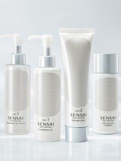 Cuatro sencillos frascos blancos de productos para el cuidado de la piel Sensai con detalles plateados, etiquetados como Paso 1 y Paso 2, dispuestos frente a un fondo suave y claro.