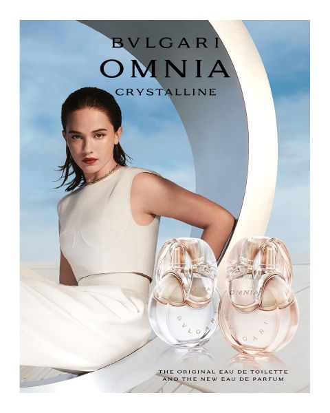 Una mujer con un vestido blanco se reclina frente a un fondo arquitectónico moderno bajo un cielo azul. En primer plano, dos frascos del perfume Bvlgari Omnia Crystalline, que muestran tonos claros y ámbar.