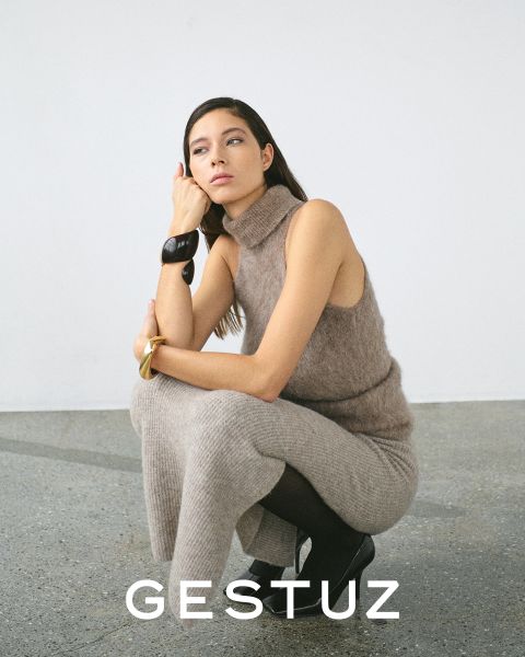 Una mujer posa con un cómodo jersey de cuello alto sin mangas de punto beige y pantalones a juego. Está en cuclillas con una rodilla levantada, lleva botas negras y un brazalete grueso. El fondo es minimalista. Abajo pone "GESTUZ".