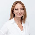 Una mujer con cabello castaño claro hasta los hombros sonríe a la cámara. Lleva una blusa blanca y un collar dorado, y está de pie frente a un fondo claro y sencillo.