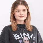Una mujer con cabello castaño hasta los hombros y una sudadera negra con un diseño gráfico está de pie frente a un fondo blanco liso. Tiene una expresión facial neutra y lleva un collar plateado.