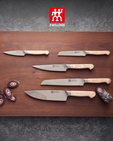 Cinco cuchillos de cocina con mangos de madera clara reposan sobre una tabla de madera, al lado hay zanahorias moradas cortadas en rodajas, y encima se ve el logo de Zwilling.