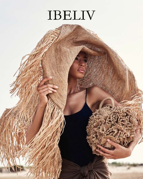 Persona con un gran sombrero de paja con flecos y un bolso estructurado a juego. Lleva un top negro sin mangas con una falda cruzada. El fondo es un exterior borroso. En la parte superior se ve la palabra "IBELIV".