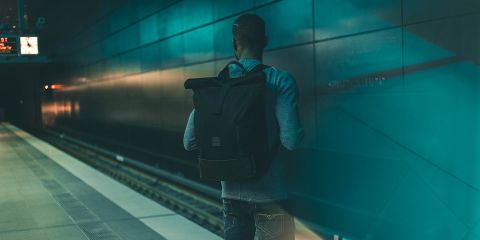 Una persona con una mochila está de pie en un andén de metro vacío, mirando las vías. La estación está tenuemente iluminada con una luz azul verdosa, creando una atmósfera evocadora. Un reloj en el techo muestra la hora, no se ve ningún tren.