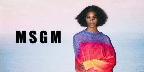 Una persona con cabello rizado está de pie junto al mar, vistiendo un colorido suéter degradado en tonos púrpura, rosa y rojo. Las letras "MSGM" son claramente visibles en el lado izquierdo de la imagen. El cielo y el mar forman un telón de fondo sereno.