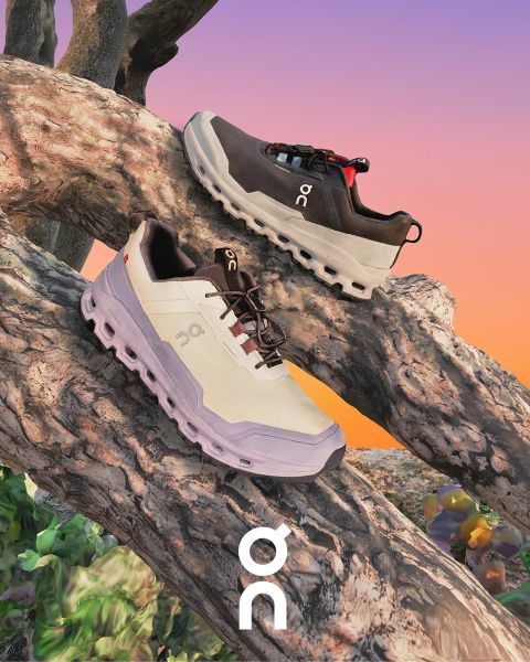 Dos zapatillas de la marca "On" se presentan sobre una rama texturizada frente a un cielo de atardecer colorido. Una zapatilla es negra con detalles grises y la otra es blanca con detalles más oscuros. Debajo, se ve el logotipo de "On".