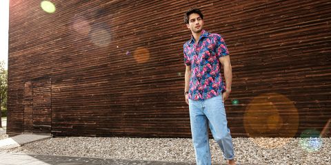 Una persona está de pie al aire libre y lleva una camisa colorida con estampado floral y vaqueros azul claro. Está de pie frente a una pared de madera, debajo de la cual hay un camino de grava. La luz del sol crea reflejos de lente en la imagen.