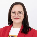 Una persona sonriente con pelo largo y oscuro y gafas, que lleva un blazer rojo sobre una camisa blanca, está de pie delante de un fondo blanco liso.