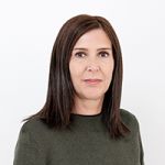 Una mujer con cabello castaño hasta los hombros y un jersey verde oscuro está de pie frente a un fondo blanco sencillo. Mira a la cámara con una expresión facial neutra.