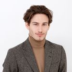 Una persona con cabello castaño y bigote lleva un blazer gris sobre un jersey de cuello alto beige. Sonríe levemente y mira a la cámara frente a un fondo blanco liso.