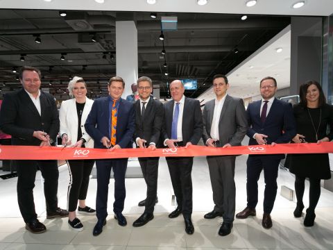 Eine Gruppe von acht Personen steht in einem Einzelhandelsgeschäft, hält große Scheren in der Hand und lächelt, während sie bereit sind, ein rotes Band mit der Aufschrift „KO“ durchzuschneiden. Sie sind in formelle Kleidung gekleidet und feiern die Eröffnung eines neuen Geschäfts.