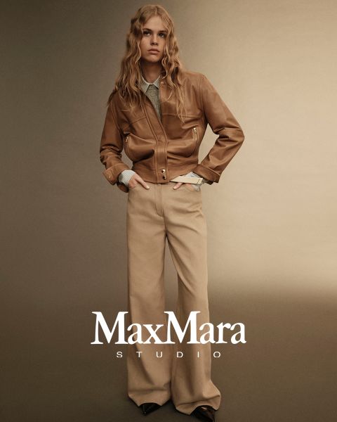 Una persona con pelo largo lleva una chaqueta de cuero marrón sobre una camisa clara y pantalones beige de pierna ancha. Están de pie frente a un fondo beige con degradado. Abajo se ve el texto "MaxMara Studio".
