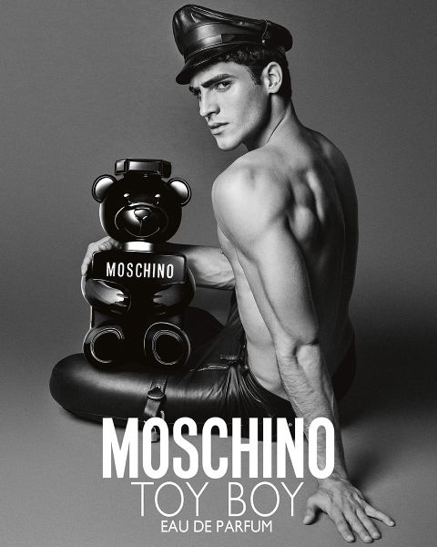 Hombre sin camisa con pantalones de cuero y sombrero sostiene un frasco de perfume Moschino con forma de oso de peluche. Texto: "MOSCHINO TOY BOY EAU DE PARFUM.
