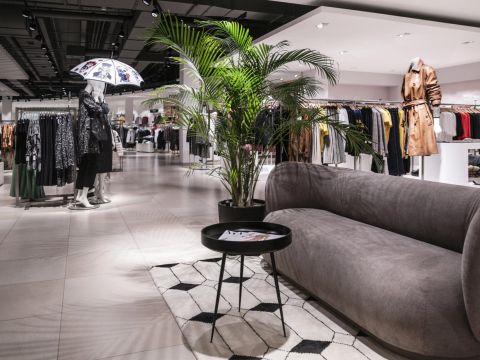 Ein modernes Interieur eines Bekleidungsgeschäfts mit einem grauen Sofa und einem runden schwarzen Tisch mit einer Pflanze auf einem geometrischen Teppich. Schaufensterpuppen präsentieren unter hellen Deckenleuchten verschiedene Outfits, darunter einen braunen Mantel und gemusterte Kleider.