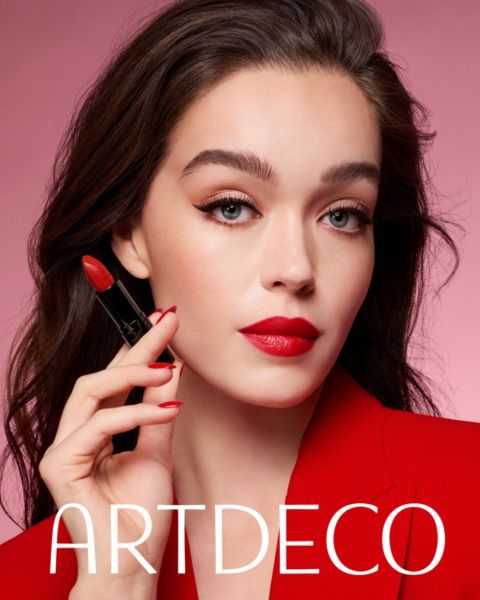 Una mujer de piel clara y cabello oscuro posa frente a un fondo rosa, sostiene un lápiz labial rojo en la mano y lleva un blazer rojo, así como un lápiz labial rojo a juego. Debajo está la inscripción "ARTDECO".