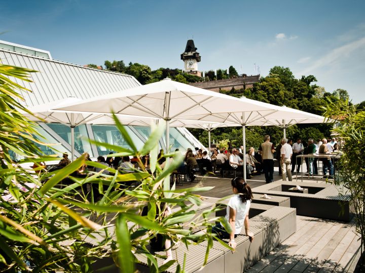 La gente disfruta de un día soleado en un café al aire libre con grandes sombrillas. En el fondo, un campanario histórico se alza sobre una colina verde. Un follaje exuberante y una arquitectura moderna rodean la zona de estar.
