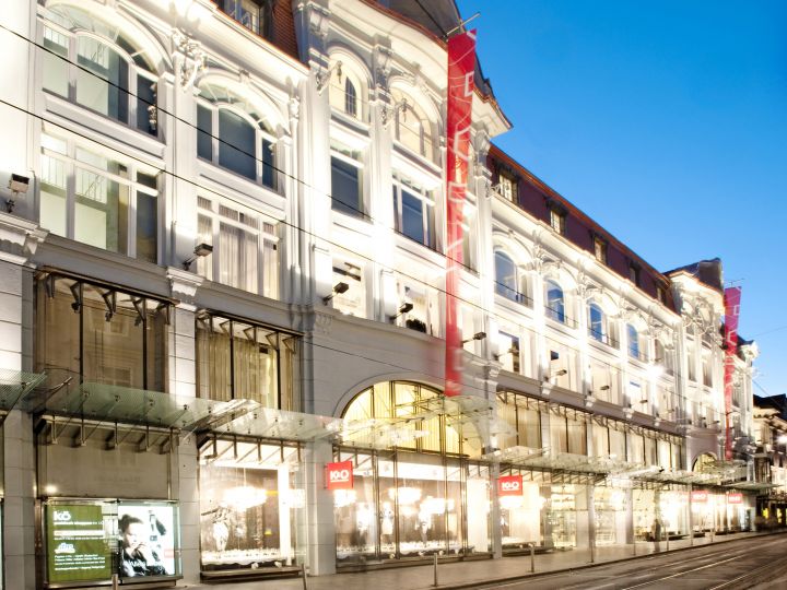 Un gran centro comercial iluminado con una fachada grandiosa, muchas ventanas arqueadas y pancartas rojas. La calle de enfrente es tranquila y el cielo es claro y azul, lo que sugiere el crepúsculo.