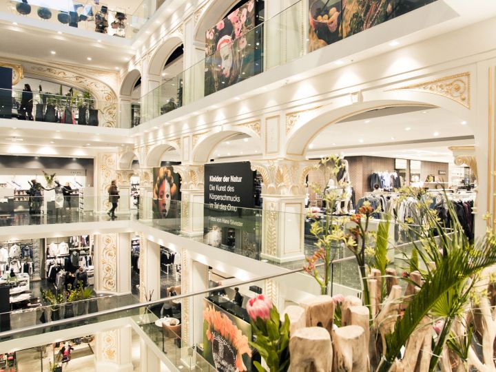 Un elegante centro comercial de varios pisos con detalles arquitectónicos clásicos. Los compradores pueden buscar ropa en tiendas con grandes ventanales. Alrededor del atrio abierto hay plantas y flores decorativas.