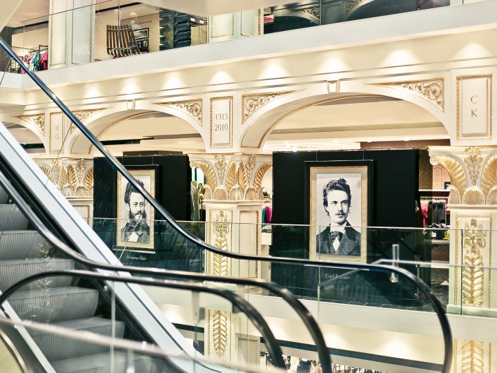 Vista interior de unos grandes almacenes ornamentados con escaleras mecánicas y dos retratos enmarcados de un hombre expuestos en el piso superior. El espacio se caracteriza por columnas decorativas e iluminación elegante.