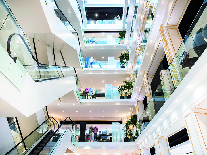 Vista interior de un moderno centro comercial de varios pisos con iluminación brillante, barandillas de vidrio, escaleras mecánicas y zonas verdes en cada planta. El diseño abierto presenta varios pisos y escaparates en un entorno espacioso.